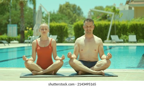 Happy Healthy Young Husband And Wife Getting Ready To Practice Yoga Together By The Pool, Sitting In Lotus Pose And Looking At Each Other Lovingly. Portrait Of A Happy Couple Relaxing On Vacation. 