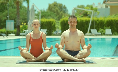 Happy Healthy Young Husband And Wife Getting Ready To Practice Yoga Together By The Pool, Sitting In Lotus Pose And Looking At Each Other Lovingly. Portrait Of A Happy Couple Relaxing On Vacation. 