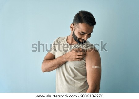 Happy healthy young Indian man showing bandage plaster on arm shoulder after getting vaccination standing on background. Vaccine and people inoculation in India, immunity for covid prevention.