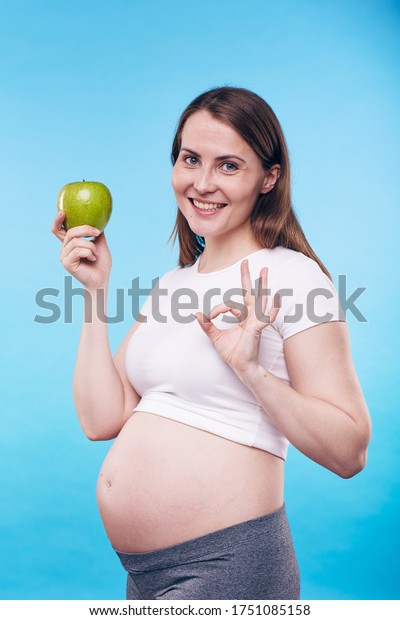 Happy\
and healthy young pregnant woman with green apple showing you okay\
gesture meaning that proper nutrition is\
important