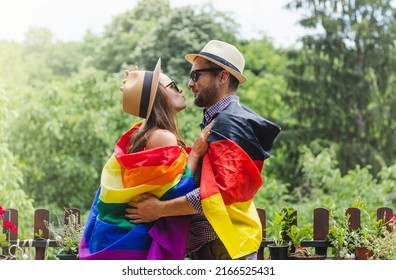 Happy Heterosexual Couple In Hats Wrapped In A German And Rainbow Flags.