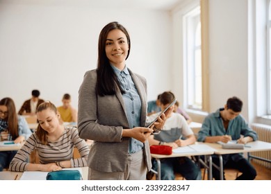 Happy high school teacher in the classroom looking at camera while her students are learning int he background. - Powered by Shutterstock