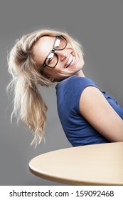 Happy high-spirited beautiful young blond woman with a beaming vivacious smile wearing glasses leaning back in her chair at a wooden table looking at the camera