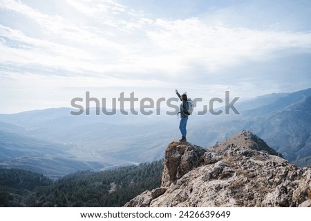 Happy hiking woman stand mountain peak looking at the view with raised hand, Young woman with backpack standing on cliff's edge and looking to a sky 