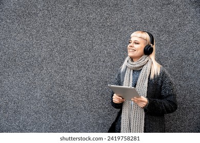 Happy hippie girl browsing a tablet and listening to the music with headphones leaning on a wall in the street. Hipster woman online working while waiting colleague listen to music on wireless headset