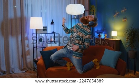 Happy hippie rocker young man in wireless headphones relaxing at home dancing on couch listening energetic disco dancing music, playing on imaginary guitar. People weekend evening leisure activities