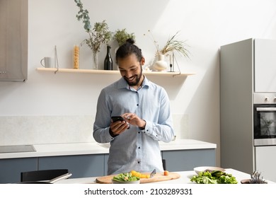 Happy Hipster Black Chef Man Using Mobile Phone In Home Kitchen, Cooking Salad, Chopping Fresh Vegetables. African American Man Consulting Organic Food Blog, Healthy Eating Recipe, Chatting Online