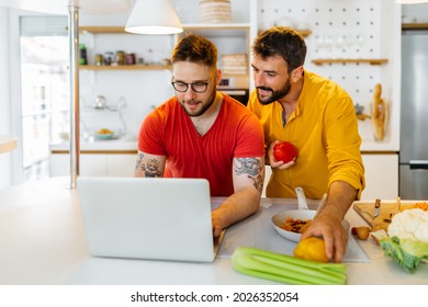 Happy Hipster Gay Couple Cooking Healthy Dinner Together. They Are Following A Recipe For A Delicious Meal On The Internet.