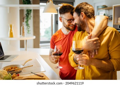 Happy hipster gay couple in love hugging while standing in the kitchen. They are cooking dinner together at home and drinking wine