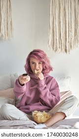 Happy Hipster Gen Z Teen Girl With Pink Hair Watching Tv Movie Series Sitting In Bed, Holding Remote Control, Eating Chips Snack In Bed. Teenager Relaxing Watching Television Reality Show In Bedroom.