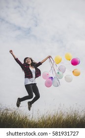 Happy hipster girl jumping with colorful toy balloons outdoors. Young woman having fun in green field against blue sky. Women freedom lifestyle concept.
