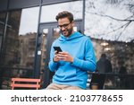 Happy hipster guy in classic spectacles for provide vision correction using cellphone gadget for checking received email message, smiling Caucasian male blogger browsing website in social media