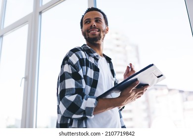 Happy hipster guy dressed in trendy shirt standing near window in coworking space spending time for creating business strategy for startup project, successful Spanish man with paperwork smiling - Shutterstock ID 2107719842