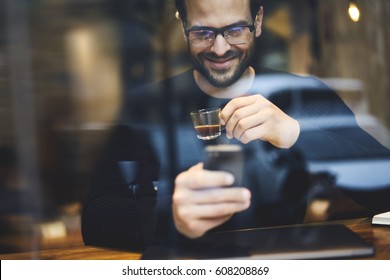 Happy Hipster Guy With Smile Looking On Smartphone Screen Watching Funny Video And Enjoying Strong Espresso In Urban Cozy Coffee Shop. Smiling Young Man Having Video Conversation With Family Members