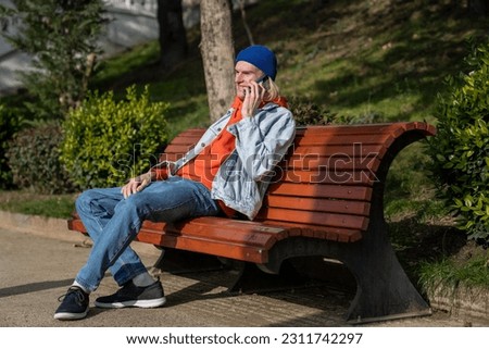 Happy hipster guy talking on mobile phone while sitting on bench in park, smiling millennial holding smartphone answering call while enjoying sunny weather outdoors, having pleasant conversation