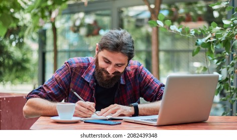 Happy Hipster Guy Writing An Essay At A Cafe. He Has His Laptop Open For Ideas And Spell Check.