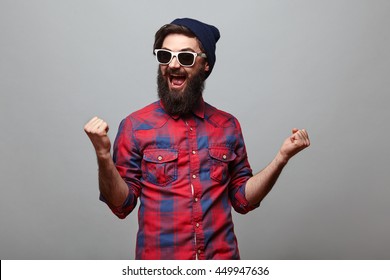 Happy Hipster Man With Beard Wearing Red Checkered Shirt And Sunglasses Exults Isolated On Grey Background