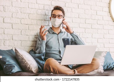 Happy Hipster Mann Freiberufler in medizinischer Gesichtsmaske sprechen Handy, sitzen auf der Couch mit Laptop auf Raps. Junge Geschäftsleute in Schutzmaske, die mit Computer im Büro arbeiten. Remote-Geschäftsbetrieb