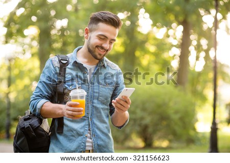 Happy hipster man walking in autumn park and smiling. Short-haited man looking at mobile phone's screen and enjoying reading messages.