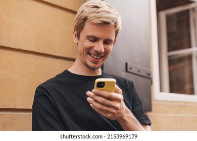 Happy Hipster Man Walking On The Street And Smiling. Short-haired Blonde Man Looking At Mobile Phone Screen And Enjoying Reading Messages. Man Texting Sms, Browsing Internet, Digital Communication.