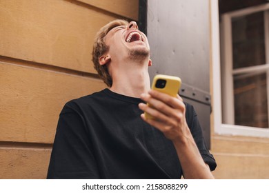 Happy hipster man walking on the street and smiling. Short-haired blonde man looking at mobile phone screen and enjoying reading messages. Man texting sms, laughing, look meme, digital communication.