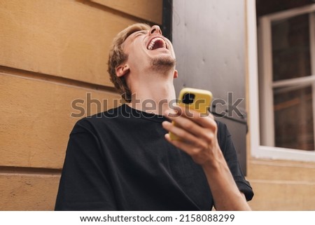 Happy hipster man walking on the street and smiling. Short-haired blonde man looking at mobile phone screen and enjoying reading messages. Man texting sms, laughing, look meme, digital communication.