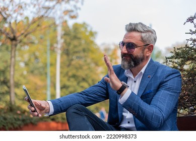 A Happy Hipster Mature Business Man Making A Video Call From His Mobile Phone  While Sitting On The Park Bench.