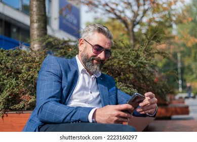 A Happy Hipster Mature Business Man Using A Mobile Phone App Texting Outside While Sitting On The Park Bench.	