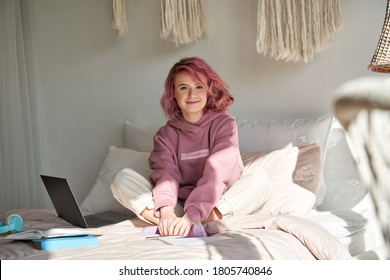 Happy hipster woman school gen z college student with pink hair wears hoodie sits on bed with laptop books learning exam looking at camera at home. Generation z ager portrait.