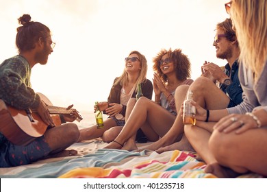 Happy hipsters relaxing and playing guitar at the beach. Friends drinking beers and listening to music. Having fun at beach party in evening.