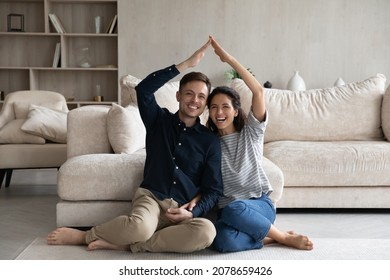 Happy Hispanic 35s Couple Joining Hands Under Heads Showing Roof Symbol Sit On Warm Floor In Fashionable Living Room. Homeowners Spouses At Relocation Day, Bank Loan For Young Family, Tenant Concept