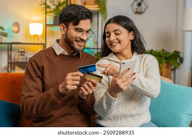 Happy Hispanic couple together doing online shopping by entering credit card number on smartphone while sitting on sofa at home. Smiling family satisfied with a good promotion on goods in apartment. - Powered by Shutterstock