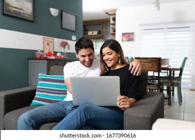 Happy Hispanic Couple Using Laptop While Sitting On Couch In Living Room At Home