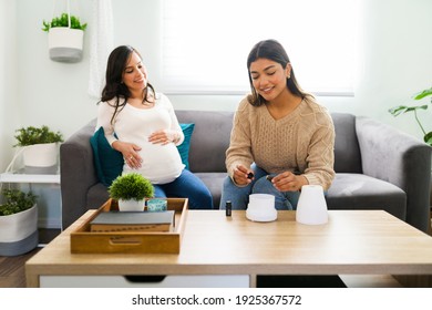 Happy hispanic doula putting essential oils with lavender in a diffuser during a home visit to help a caucasian pregnant woman to rest better and relax 
