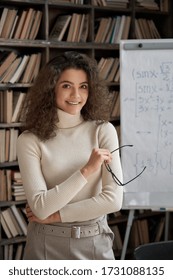 Happy hispanic ethnic young woman university teacher, smiling latin girl college student, professional tutor holding glasses looking at camera standing in classroom with whiteboard. Vertical portrait.