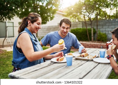 Happy Hispanic Family Eating Grilled Hot Dogs On Picnic Table In Backyard