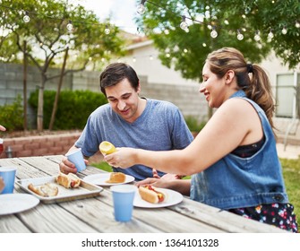 Happy Hispanic Family Eating Grilled Hot Dogs On Picnic Table In Backyard