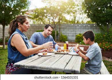 Happy Hispanic Family Eating Grilled Hot Dogs On Picnic Table In Backyard