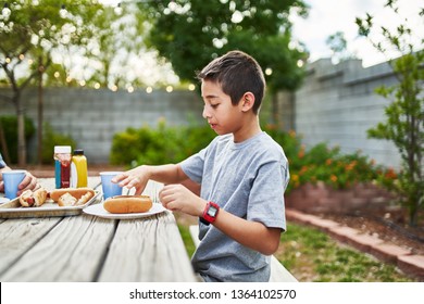 Happy Hispanic Family Eating Grilled Hot Dogs On Picnic Table In Backyard