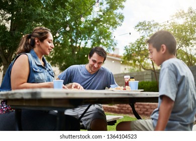 Happy Hispanic Family Eating Grilled Hot Dogs On Picnic Table In Backyard