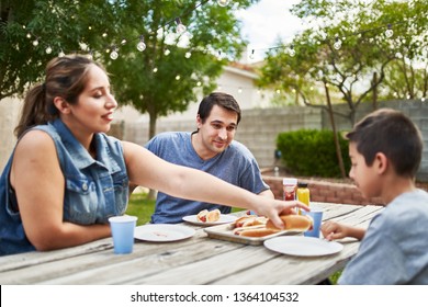 Happy Hispanic Family Eating Grilled Hot Dogs On Picnic Table In Backyard