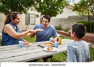 Happy Hispanic Family Eating Grilled Hot Dogs On Picnic Table In Backyard