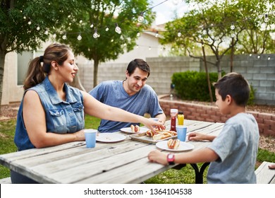 Happy Hispanic Family Eating Grilled Hot Dogs On Picnic Table In Backyard