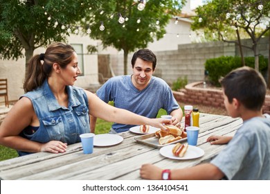 Happy Hispanic Family Eating Grilled Hot Dogs On Picnic Table In Backyard