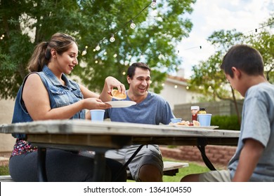 Happy Hispanic Family Eating Grilled Hot Dogs On Picnic Table In Backyard