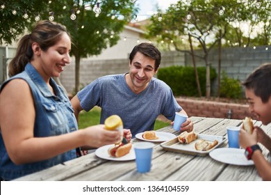 Happy Hispanic Family Eating Grilled Hot Dogs On Picnic Table In Backyard