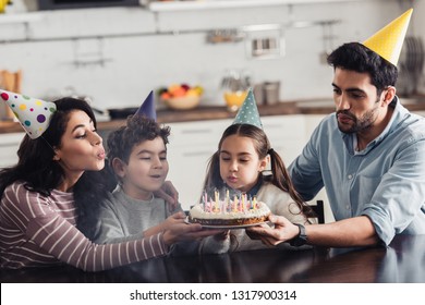 Happy Hispanic Family In Party Caps Blowing At Candles On Birthday Cake At Home