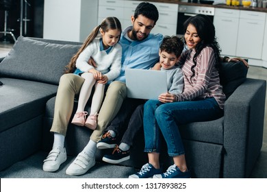 Happy Hispanic Family Sitting On Sofa And Looking At Laptop At Home