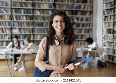 Happy Hispanic gen Z student girl with headphones visiting public library for work on study research project, holding learning papers, notebook, looking at camera, smiling. Head shot portrait - Shutterstock ID 2111421341