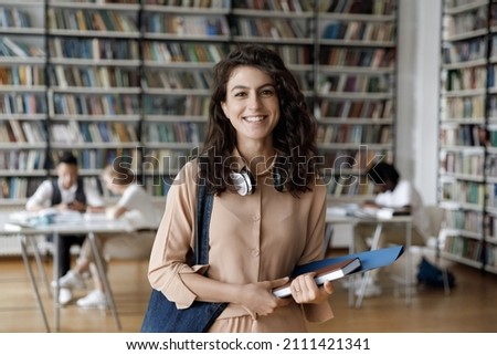 Happy Hispanic gen Z student girl with headphones visiting public library for work on study research project, holding learning papers, notebook, looking at camera, smiling. Head shot portrait